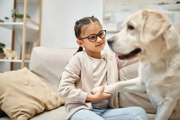 Glückliches Mädchen im Grundalter mit Brille, die Pfote eines Labradors haltend und auf dem Sofa im Wohnzimmer sitzend — Stockfoto
