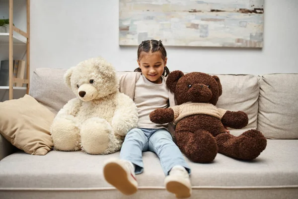 Bonito elementar idade criança no casual desgaste sentado no sofá com macio ursinhos de pelúcia na sala de estar — Fotografia de Stock