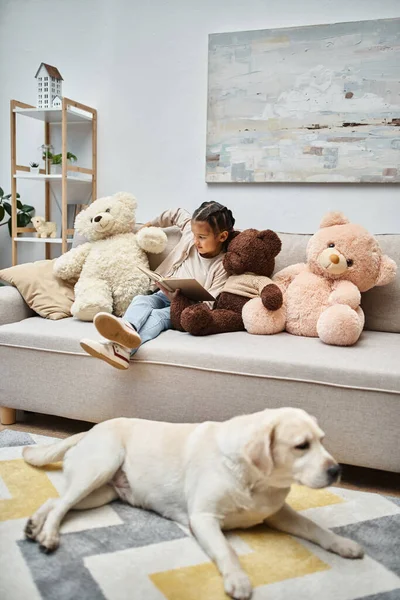 Adorable chica sentada en sofá con suaves osos de peluche y libro de lectura cerca de labrador en sala de estar - foto de stock