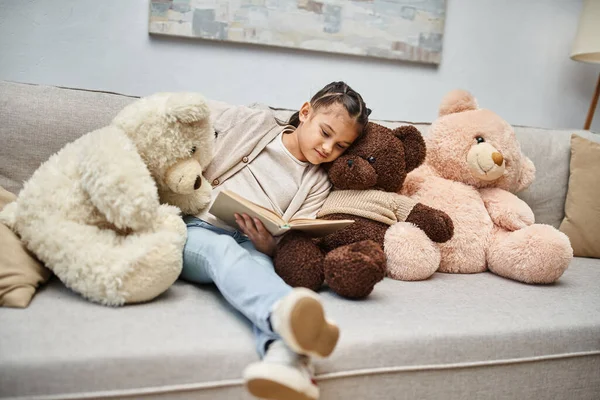 Adorable chica sentada en el sofá con suaves osos de peluche y libro de lectura en la sala de estar moderna - foto de stock
