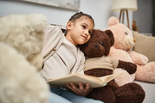 Cute girl sitting on comfortable sofa with soft teddy bears and reading book in modern living room — Stock Photo