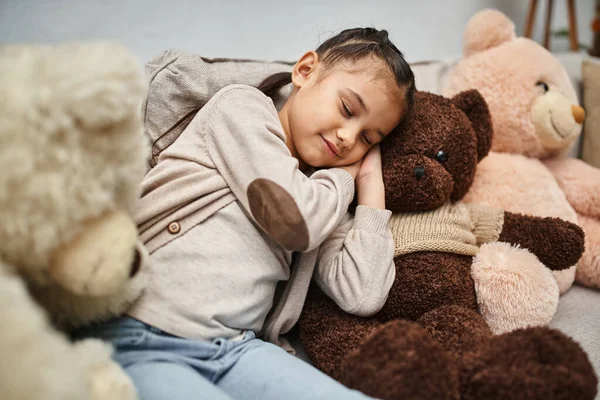 Adorable chica de edad elemental durmiendo entre suaves osos de peluche en el sofá en la sala de estar moderna - foto de stock