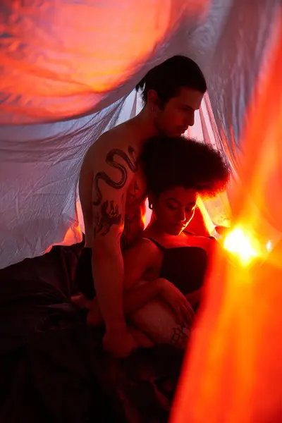 Couple multiculturel sexy assis sous le drap de lit et posant amoureusement entouré de lumières, ensemble — Photo de stock
