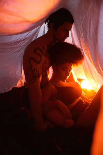 Young diverse couple sitting under bedsheet and posing lovingly surrounded by lights, togetherness — Stock Photo