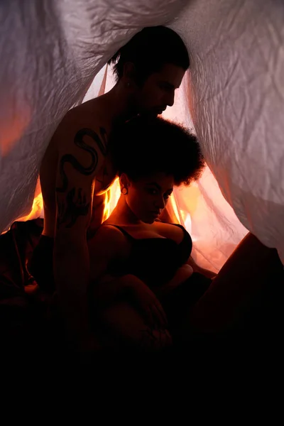 Jeune couple multiculturel assis sous le drap de lit et posant entouré de lumières alléchantes — Photo de stock