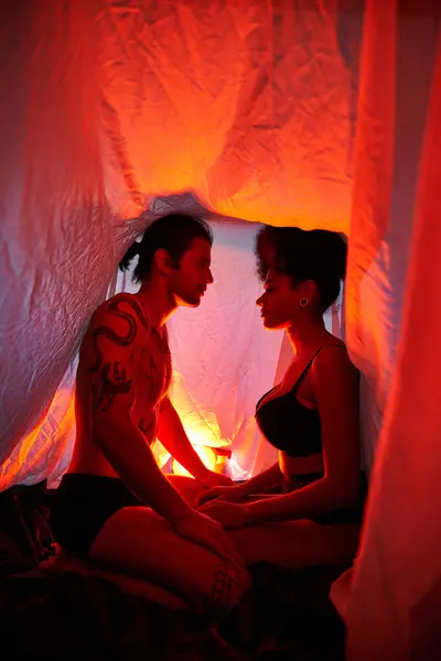 Sexy diverse couple sitting under bedsheet and posing alluringly surrounded by lights, together — Stock Photo