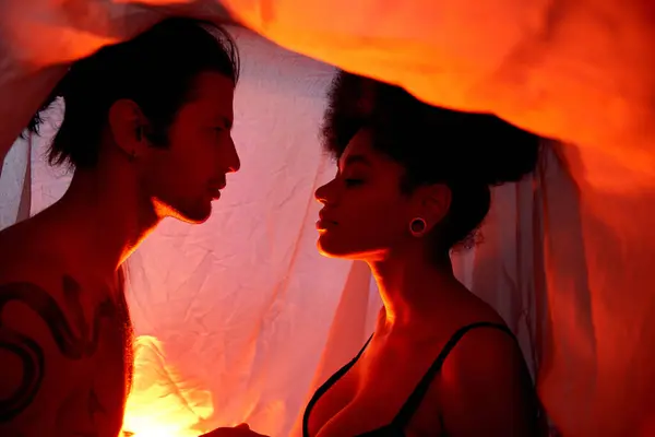 Tempting diverse couple sitting under bedsheet and posing surrounded by lights, togetherness — Stock Photo