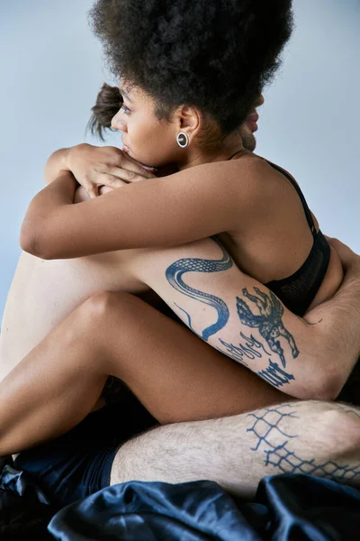 Beautiful young multicultural couple sitting on their bed and hugging lovingly on gray backdrop — Stock Photo