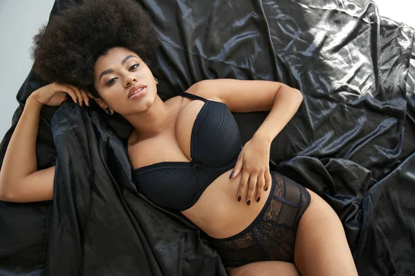 Вид сверху на сексуальную африканскую женщину в черном нижнем белье, лежащую в постели и смотрящую в камеру — стоковое фото