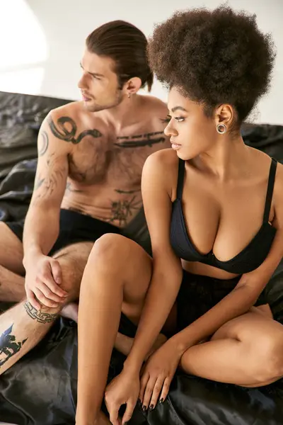 Homme attrayant avec des tatouages assis sur le lit à côté de sa petite amie afro-américaine, couple sexy — Photo de stock