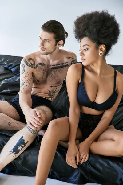 Homem bonito com tatuagens sentado na cama ao lado de sua namorada americana africana bonita, casal sexy — Fotografia de Stock