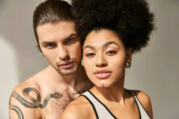 Handsome man with tattoos hugging lovingly his pretty african american girlfriend on gray backdrop — Stock Photo