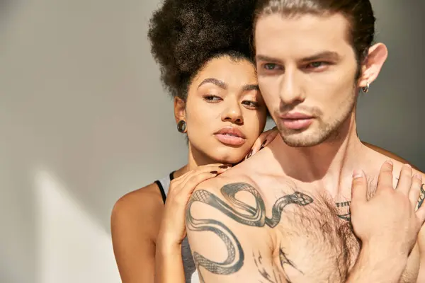 Sexy junger Mann mit Tätowierungen umarmt seine schöne afrikanisch-amerikanische Freundin vor grauem Hintergrund — Stockfoto