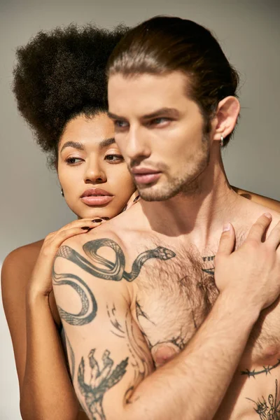 Alluring man with tattoos hugging lovingly his pretty african american girlfriend on gray backdrop — Stock Photo
