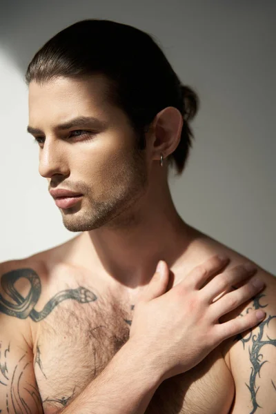Appealing young man with tattoos and ponytail posing with his hand on chest and looking away — Stock Photo