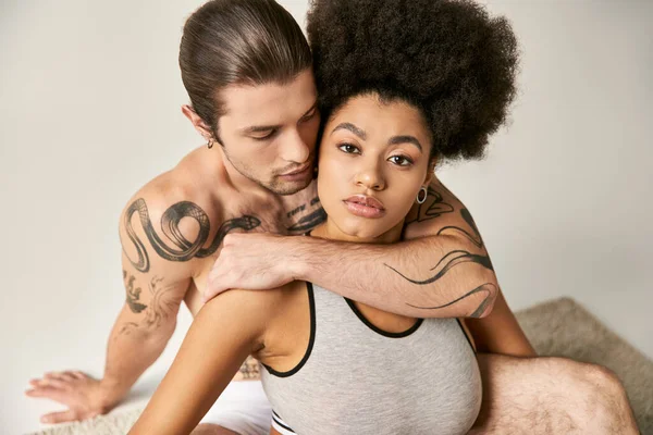 Joven hombre tatuado abrazando a su bonita novia afroamericana en gris telón de fondo, sexy pareja - foto de stock