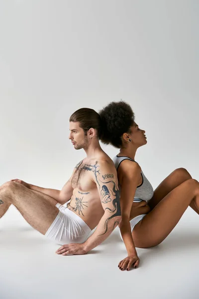 Appealing young diverse boyfriend and girlfriend sitting back to back on floor, sexy couple — Stock Photo