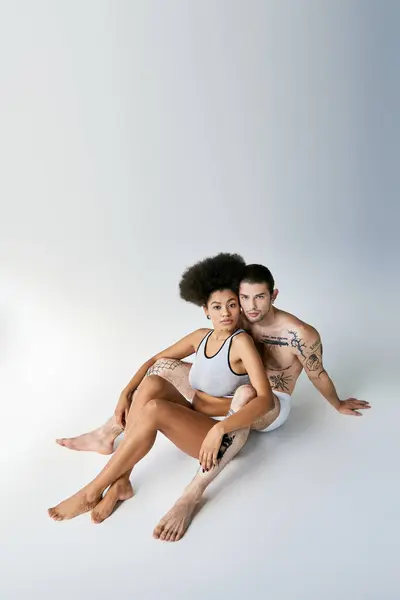 Bien parecido sexy pareja multicultural en ropa interior cómoda sentado en el suelo y abrazándose calurosamente - foto de stock