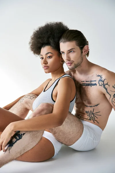 Atractiva pareja multicultural sexy en ropa interior cómoda sentada en el suelo y abrazándose amorosamente - foto de stock