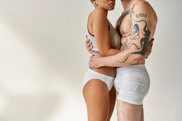 Обрезанный вид мультикультурной пары в нижнем белье позирует вместе и обнимает тепло друг друга — стоковое фото