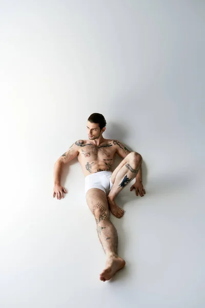 Вид зверху добре виглядає молодий чоловік з хвостом і татуюваннями в нижній білизні, що лежить на підлозі привабливо — стокове фото