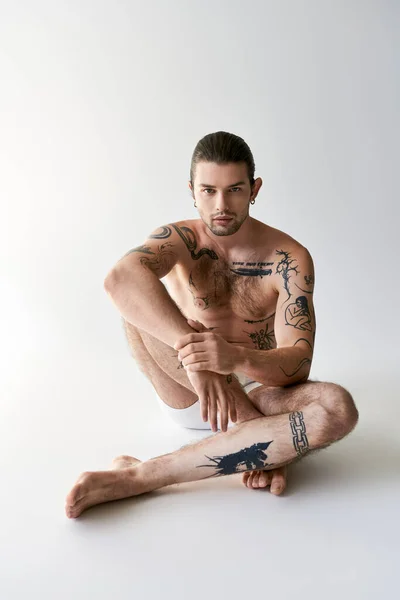 Boa aparência jovem tatuado modelo masculino em roupa interior sentado no chão e olhando para a câmera — Stock Photo