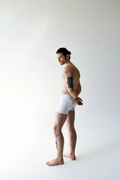 Sexy strong man with ponytail and cool tattoos posing in comfy underwear on ecru background — стоковое фото