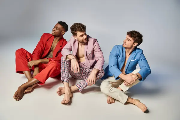 Cheerful multiracial friends in vibrant colorful suits sitting on floor with crossed legs, fashion — Stock Photo