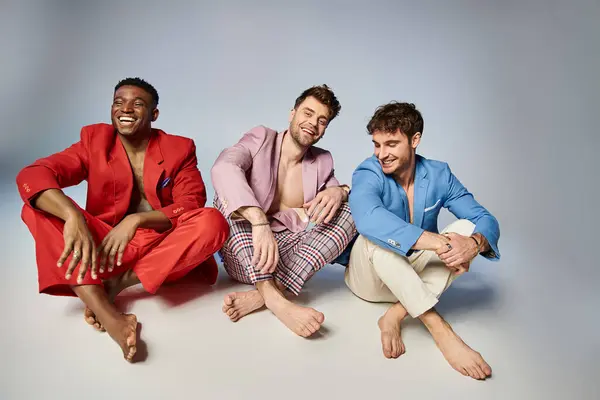 Fröhliche multiethnische Männer in hellen Anzügen, die mit überkreuzten Beinen auf dem Boden sitzen und freudig lächeln — Stockfoto