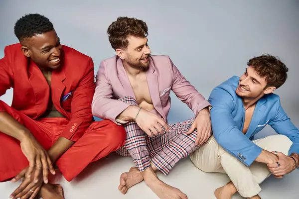 Homens diversos alegres em ternos sentados no chão com pernas cruzadas e sorrindo alegremente, moda — Fotografia de Stock