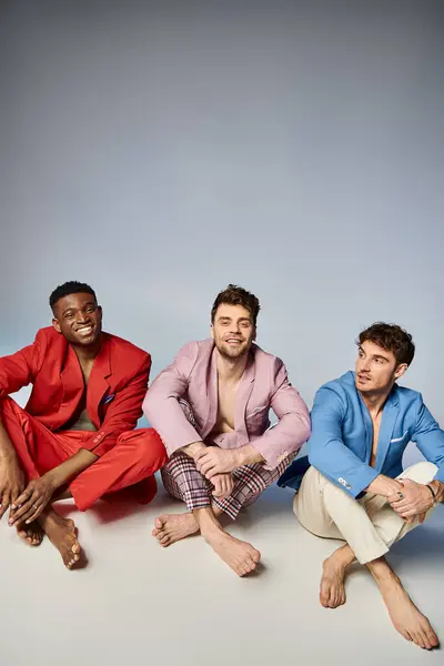 Glücklich diverse Männer in lebendigen Anzügen, die mit überkreuzten Beinen auf dem Boden sitzen und fröhlich lächeln, Mode — Stockfoto
