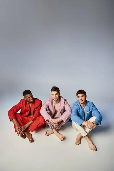 Щасливі міжрасові чоловіки в яскравих костюмах сидять на підлозі з схрещеними ногами і радісно посміхаються — стокове фото