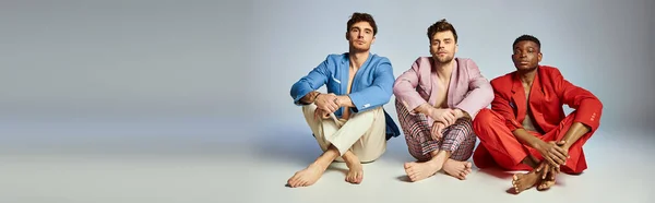 Três homens bonitos e diversos em vibrantes ternos da moda sentados no chão com pernas cruzadas, banner — Fotografia de Stock