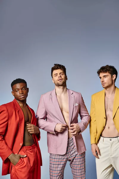 Três homens diversos em ternos desabotoados vibrantes posando juntos em pano de fundo cinza, conceito de moda — Fotografia de Stock