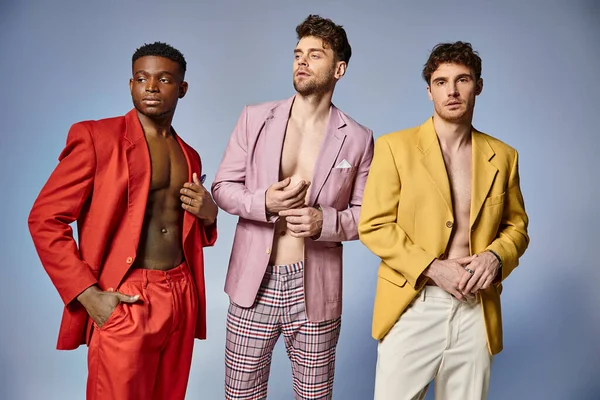 Appealing interracial men in unbuttoned vibrant suits posing on gray backdrop, fashion concept — Stock Photo