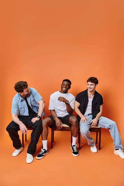 Homens inter-raciais alegres em roupas urbanas casuais sorrindo alegremente em pano de fundo laranja, conceito de moda — Fotografia de Stock