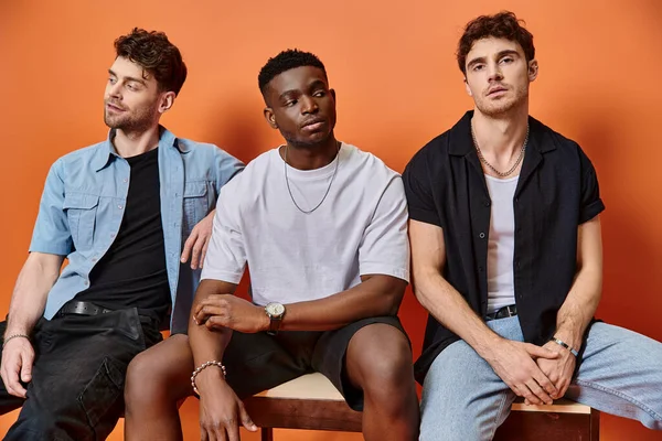 Attractive multicultural male models in casual urban attires sitting on chairs on orange backdrop — Stock Photo