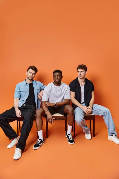 Good looking multicultural male models in casual urban attires sitting on chairs on orange backdrop — Stock Photo