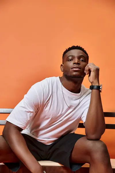 Good looking young african american man in urban outfit sitting on bench on orange backdrop, fashion — Stock Photo