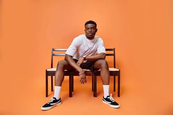 Attrayant jeune homme afro-américain en tenue urbaine assis sur un banc sur fond orange, la mode — Photo de stock