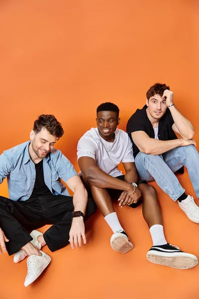Trois hommes multiraciaux joyeux en vêtements urbains décontractés souriants et assis sur le sol, concept de mode — Photo de stock