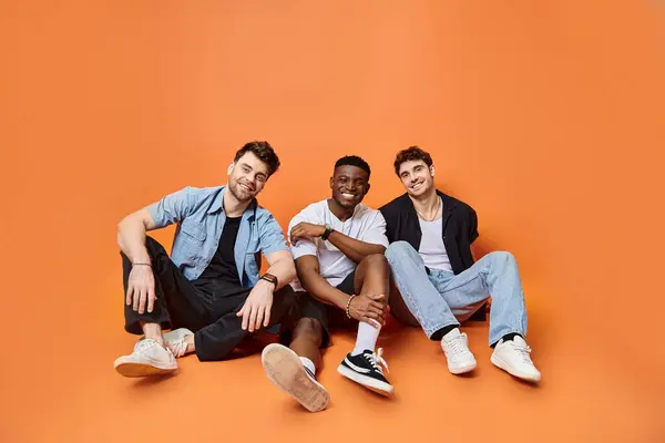 Trois hommes joyeux et multiethniques en vêtements urbains décontractés souriants et assis sur le sol, concept de mode — Photo de stock