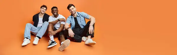 Trois hommes heureux divers en vêtements décontractés souriant et assis sur le sol, concept de mode, bannière — Photo de stock