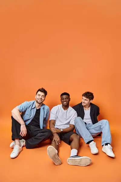 Fröhliche multirassische Freunde in lässigen Straßenoutfits, die auf dem Boden sitzen und glücklich lächeln, Mode — Stockfoto