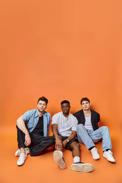 Séduisants hommes élégants multiculturels dans des tenues urbaines à la mode assis sur le sol, concept de mode — Photo de stock