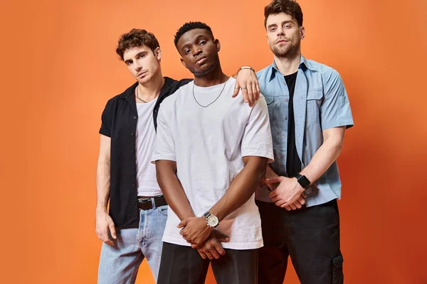Appealing multiracial male models in street attires looking at camera on orange backdrop, fashion — Stock Photo