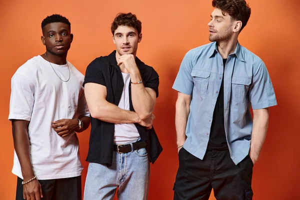 Modelos masculinos multirraciais de boa aparência em roupas urbanas elegantes posando em pano de fundo laranja, moda — Fotografia de Stock