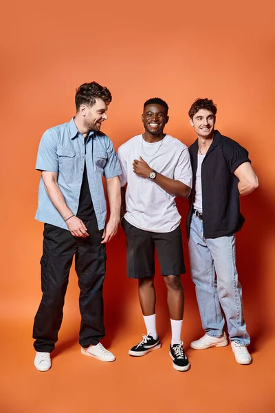 Joyful multicultural men in casual street wear posing happily together on orange backdrop, fashion — Stock Photo