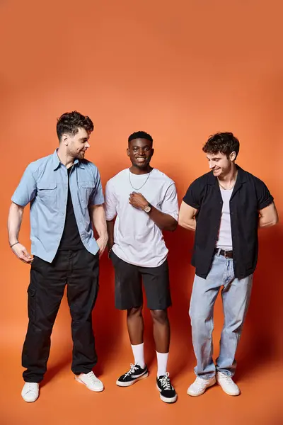 Alegre multicultural hombres en casual calle desgaste posando felizmente juntos en naranja telón de fondo, moda - foto de stock