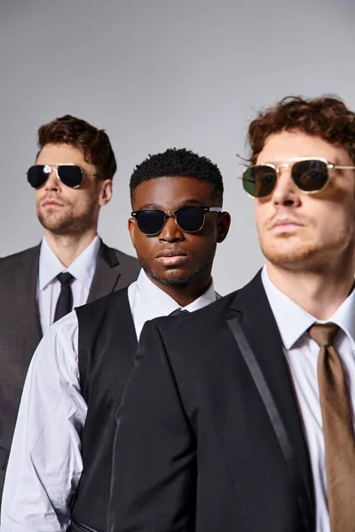 Handsome multiracial male models in elegant smart suits with sunglasses posing on gray backdrop — Stock Photo